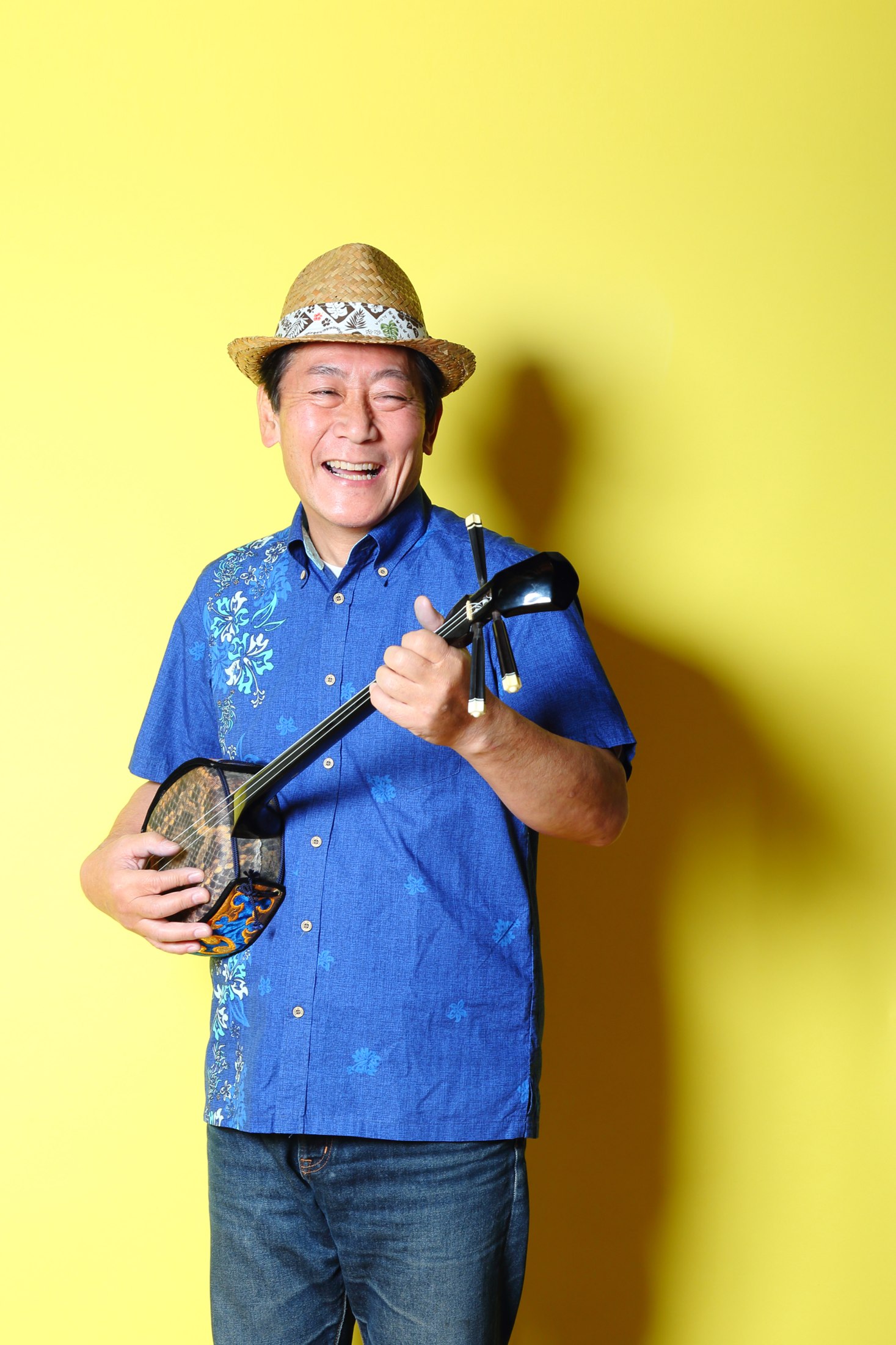 黄色い背景で、帽子と青いシャツを身につけ弦楽器を持つ男性